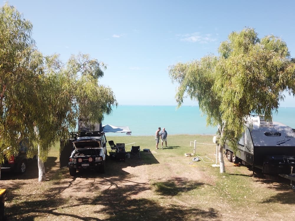Photo of a two 4WDs parked on the left and right of the screen with 2 men having a conversation in the center of the image with a beach in the background.
