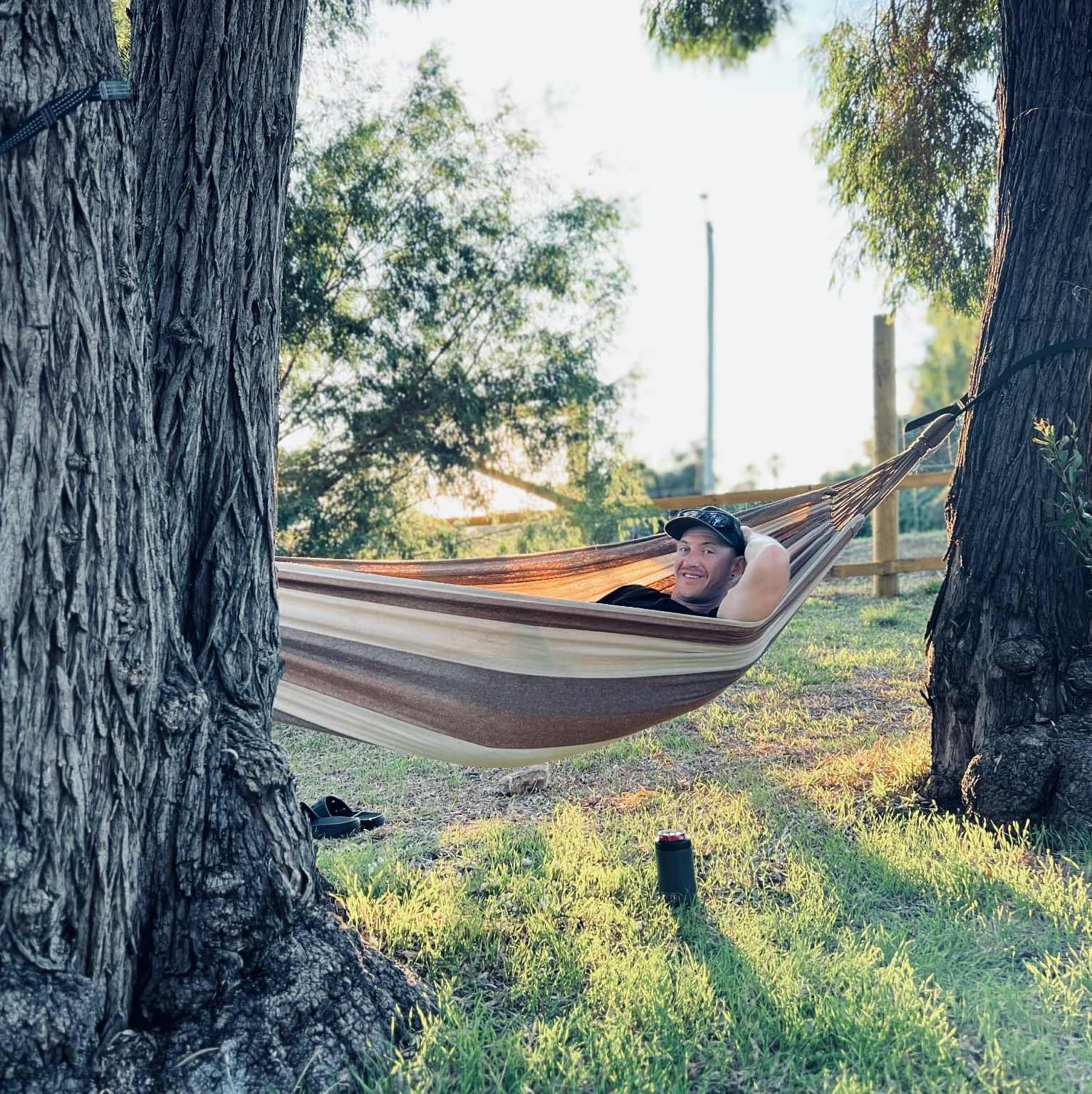 A man called Combi laying in a brown and tan stripd hammock between two australian native trees.