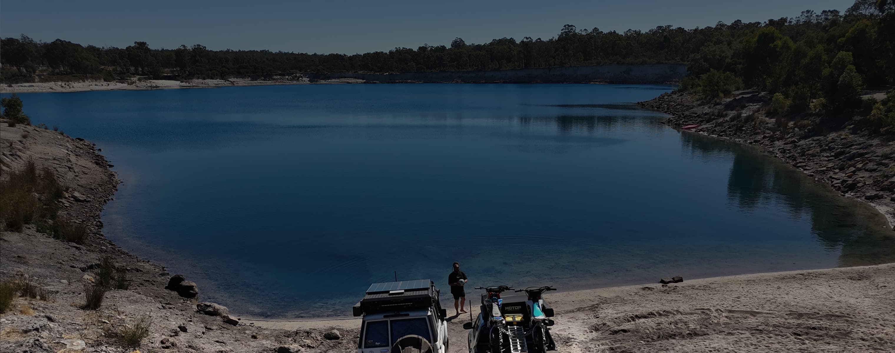 Two off-road vehicles parked by a blue lake surrounded by rocky terrain and forest at dusk.